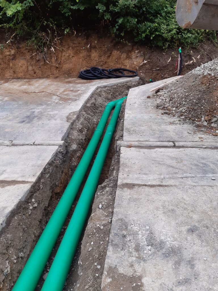 Zwei grüne Kunststoffrohre, die in einem frisch ausgehobenen Graben über einer Betonfläche verlegt sind.