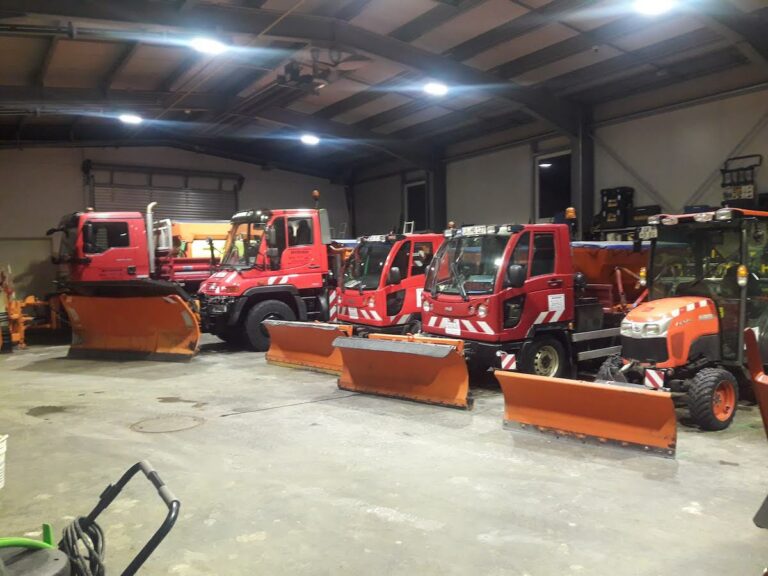 Mehrere rote Winterdienstfahrzeuge mit Schneeschilden stehen in einer Halle nebeneinander, bereit für den Einsatz bei Schnee und Eis.
